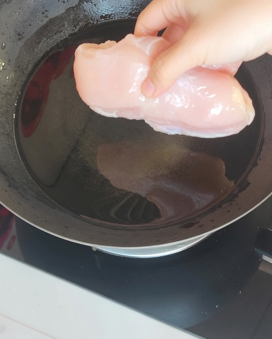 Pechuga de pollo sumergida en agua hirviendo dentro de un wok junto con aromáticos como jengibre y cebolleta.