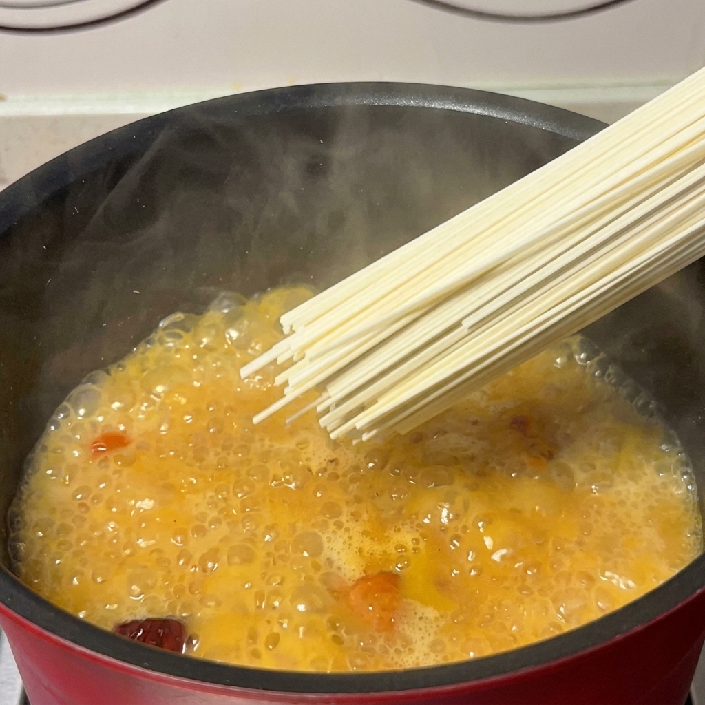 Un puñado de fideos secos introduciéndose en una olla con caldo dorado de pollo y setas.