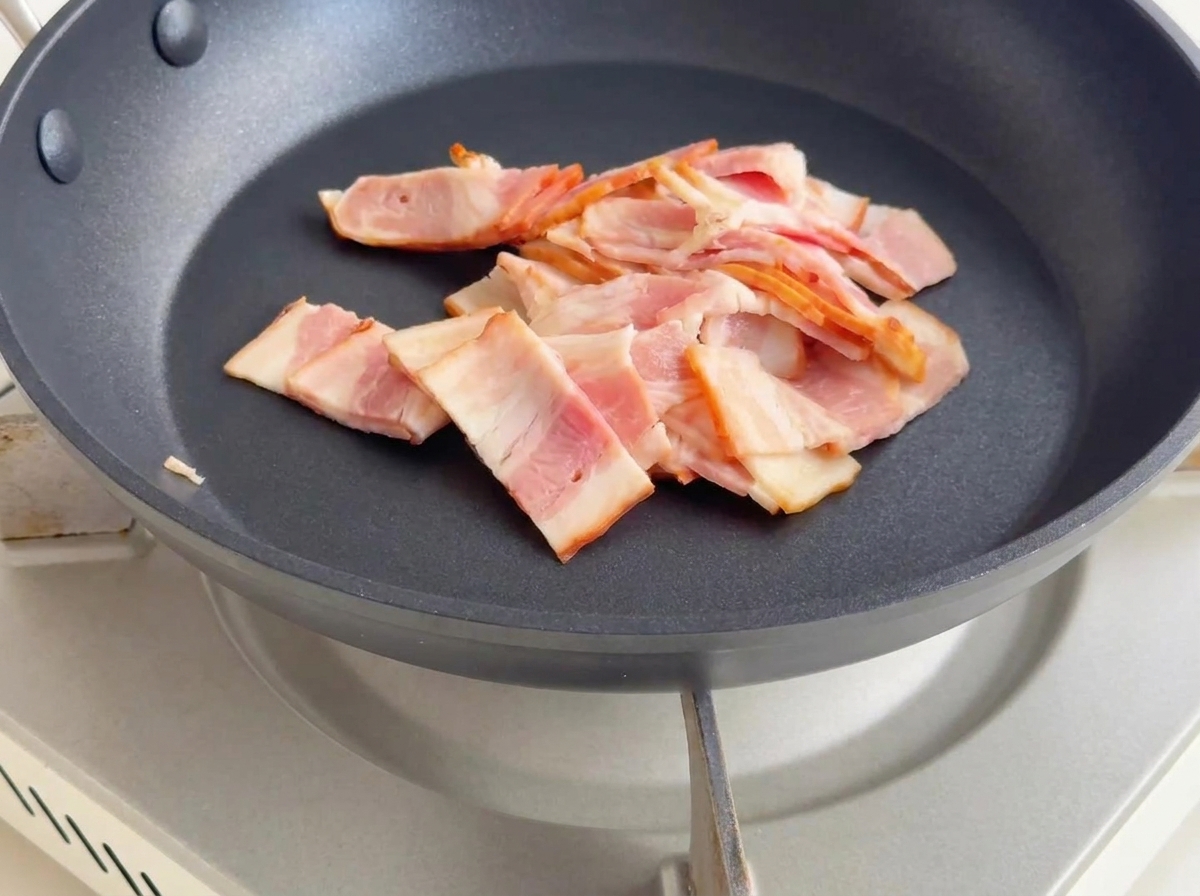 Trozos de tocino friéndose en una sartén, dorándose y soltando grasa.