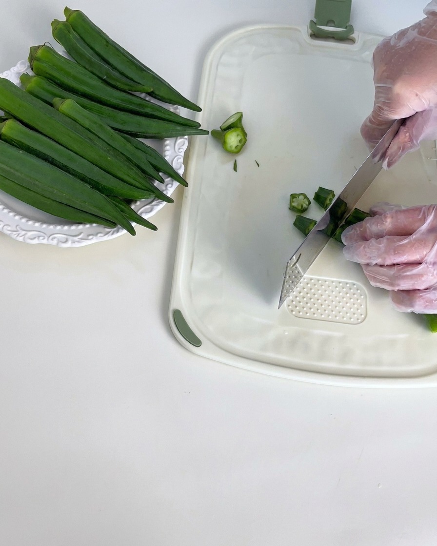 Vainas de okra verde fresca siendo recortadas en una tabla de cortar blanca.