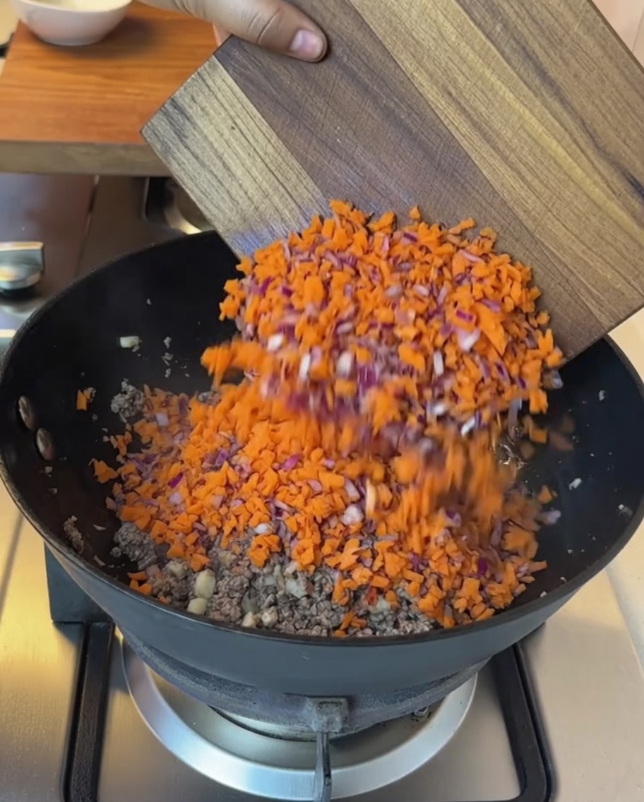 Verduras picadas y tomates troceados siendo añadidos a la sartén con la carne cocida.
