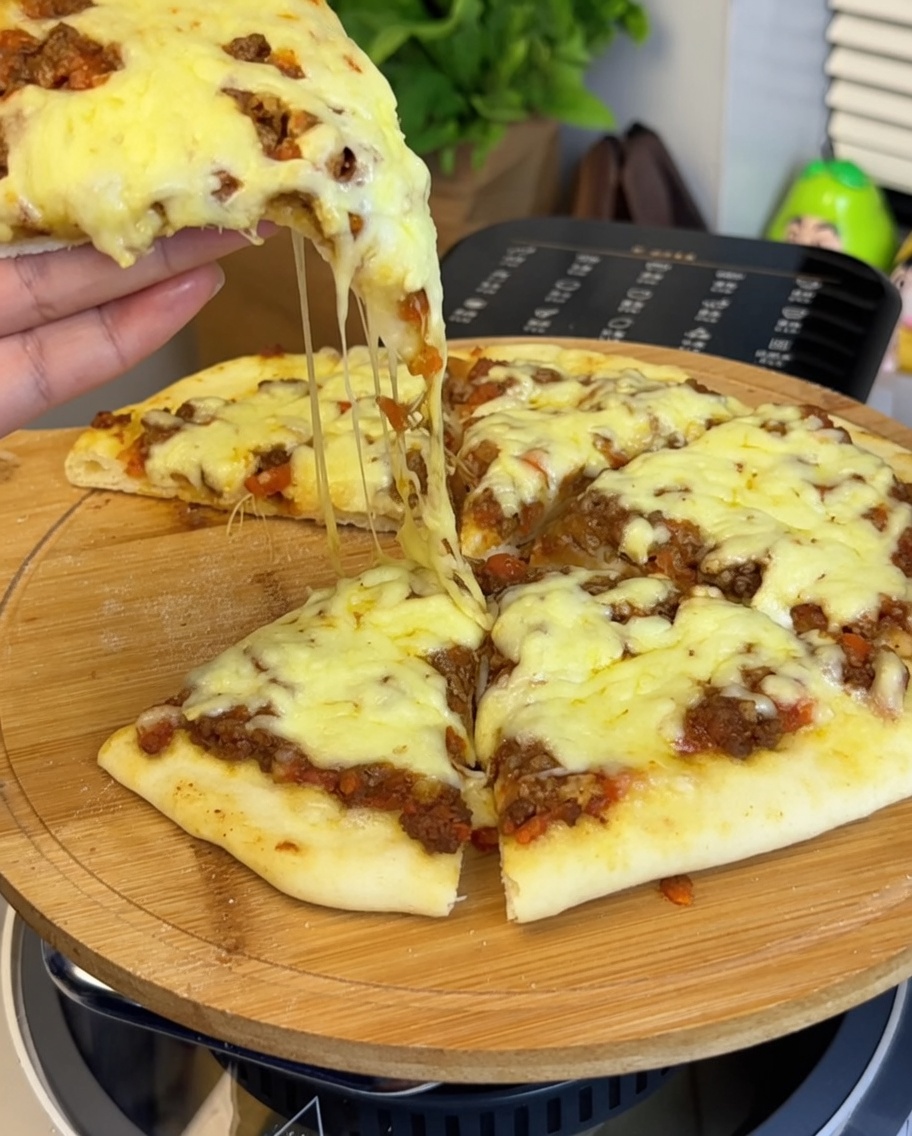 Primer plano de la salsa de carne extendiéndose sobre la masa, con una mano sosteniendo un trozo de pizza mostrando queso derretido.