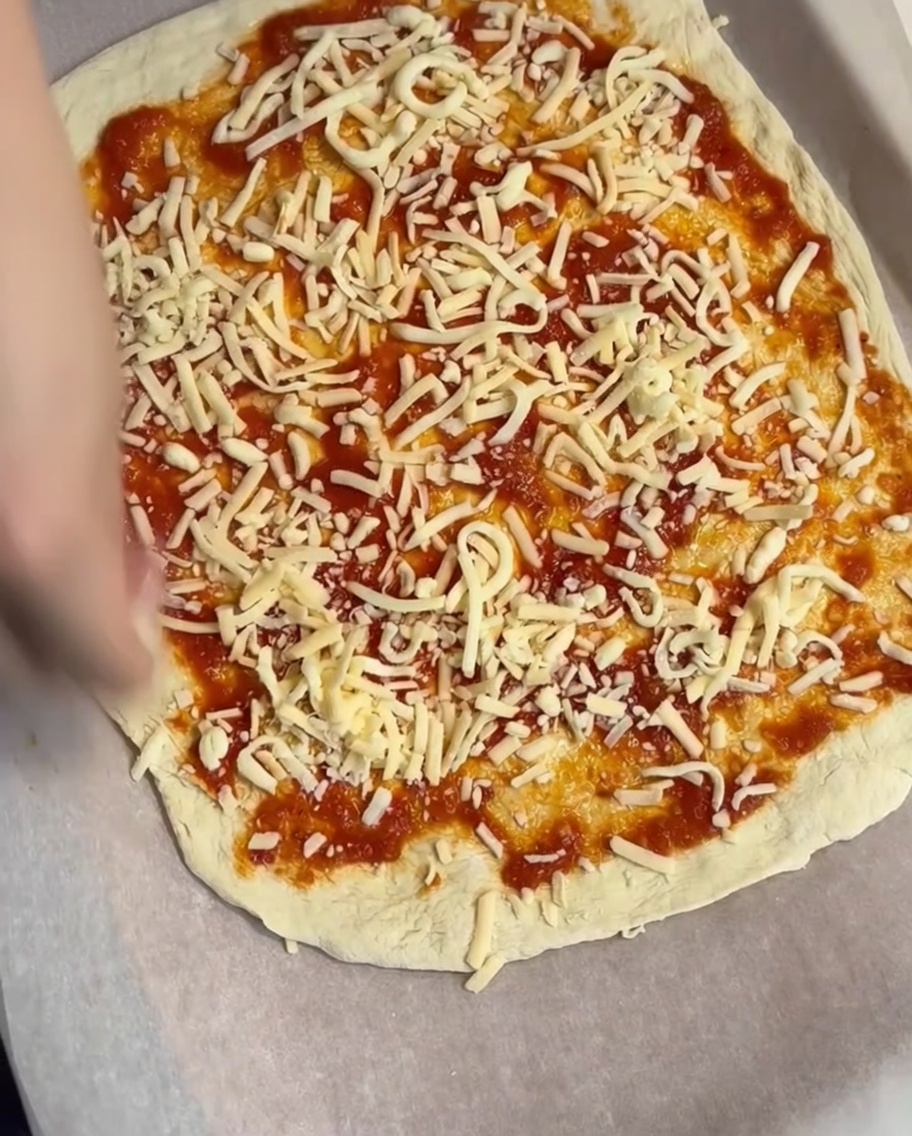 Capa de queso rallado siendo esparcida sobre la base de salsa de tomate.