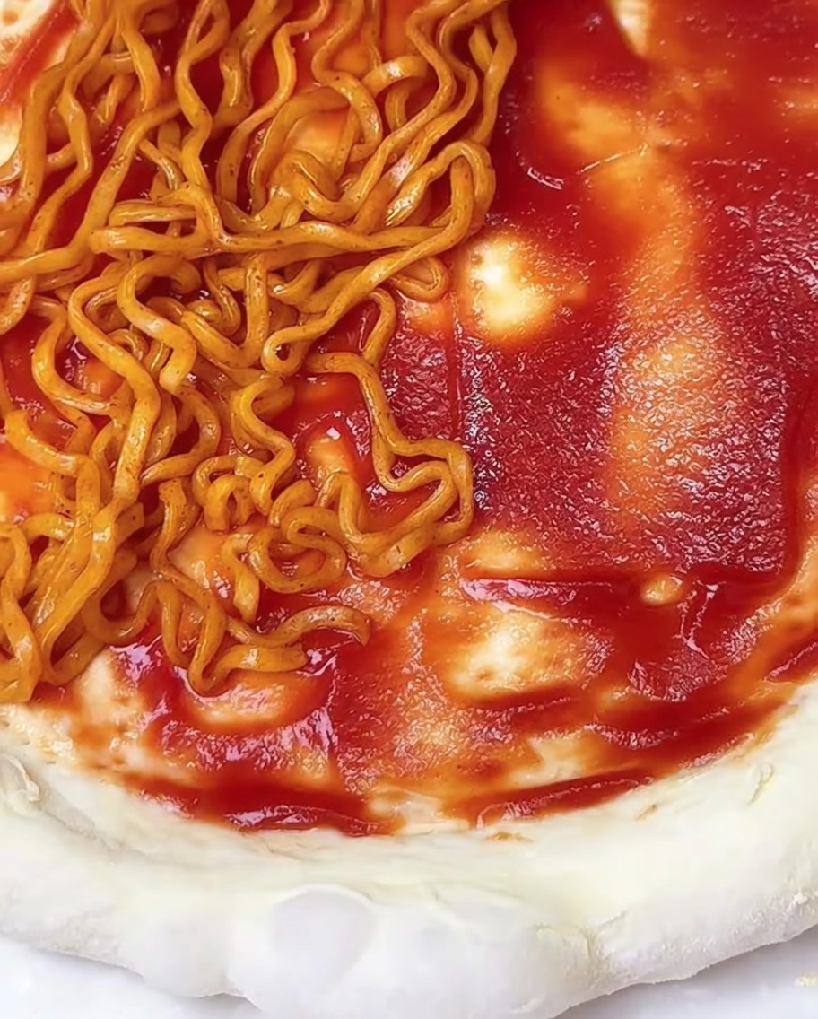 Un primer plano de una masa de pizza plana untada con salsa de tomate roja, cubierta parcialmente con fideos ramen picantes y copos de algas.