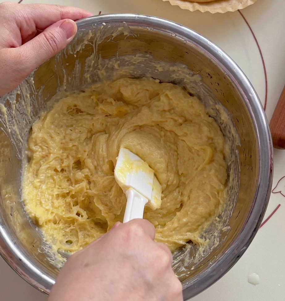Primer plano de una espátula blanca machacando durián amarillo en un bol metálico.