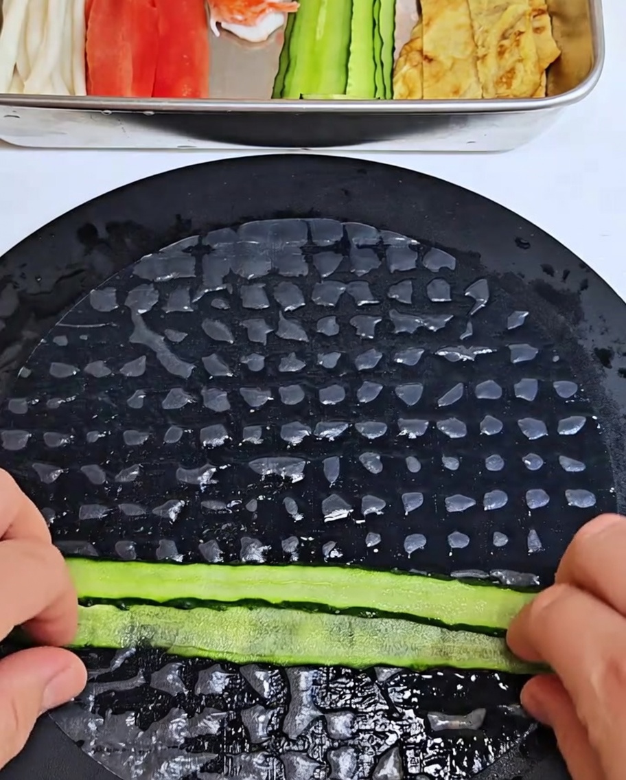 Tiras de pepino siendo colocadas sobre papel de arroz suavizado y enrolladas en un cilindro apretado.