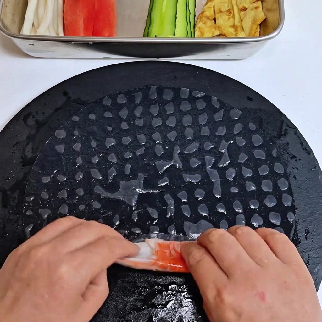 Tiras de palitos de surimi siendo envueltas y enrolladas en una lámina de papel de arroz suavizado.