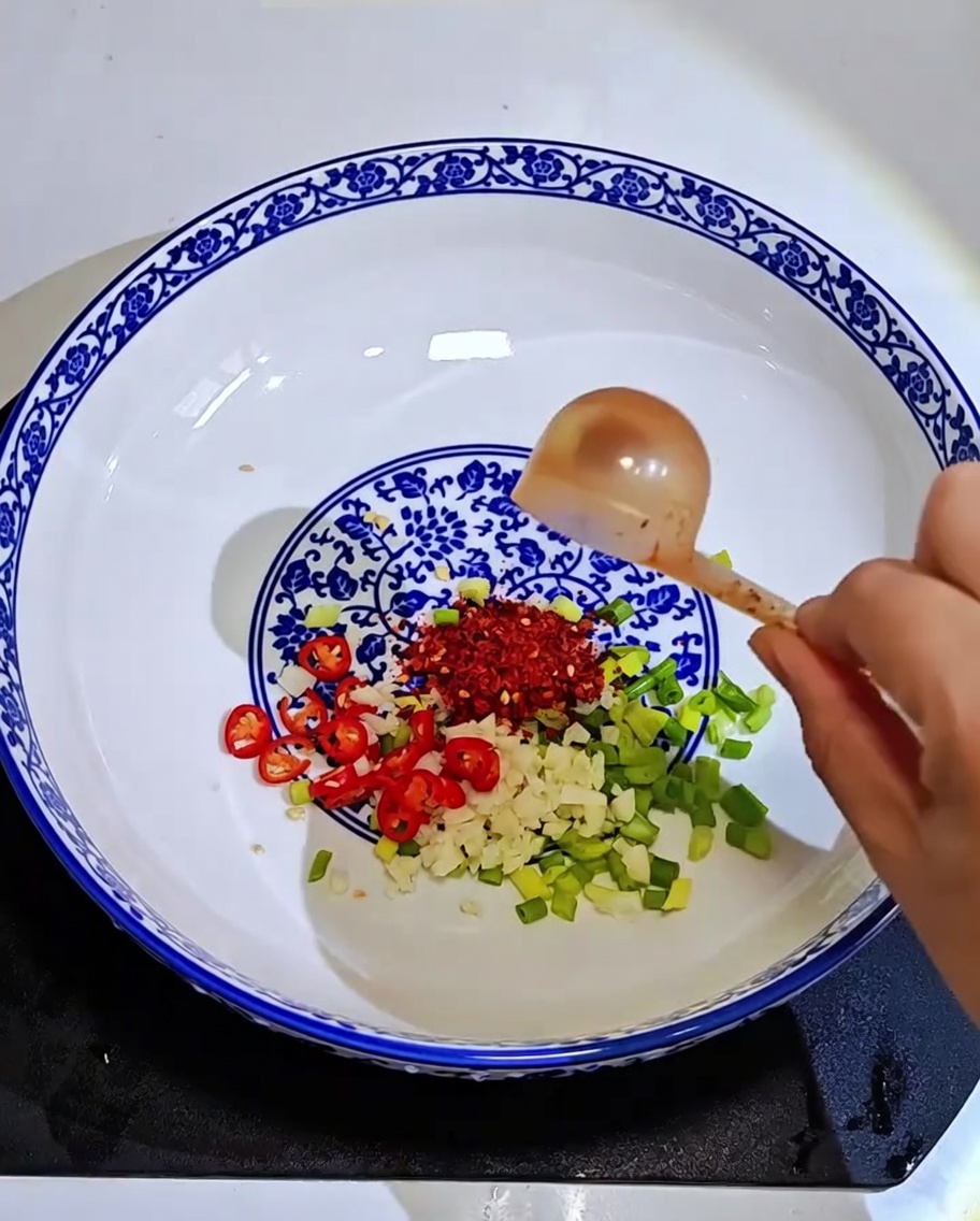 Primer plano de un tazón que contiene ajo, cebollino y copos de chile siendo rociados con aceite caliente.