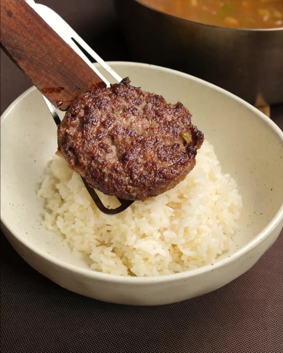 Hamburguesa siendo colocada sobre un tazón de arroz.