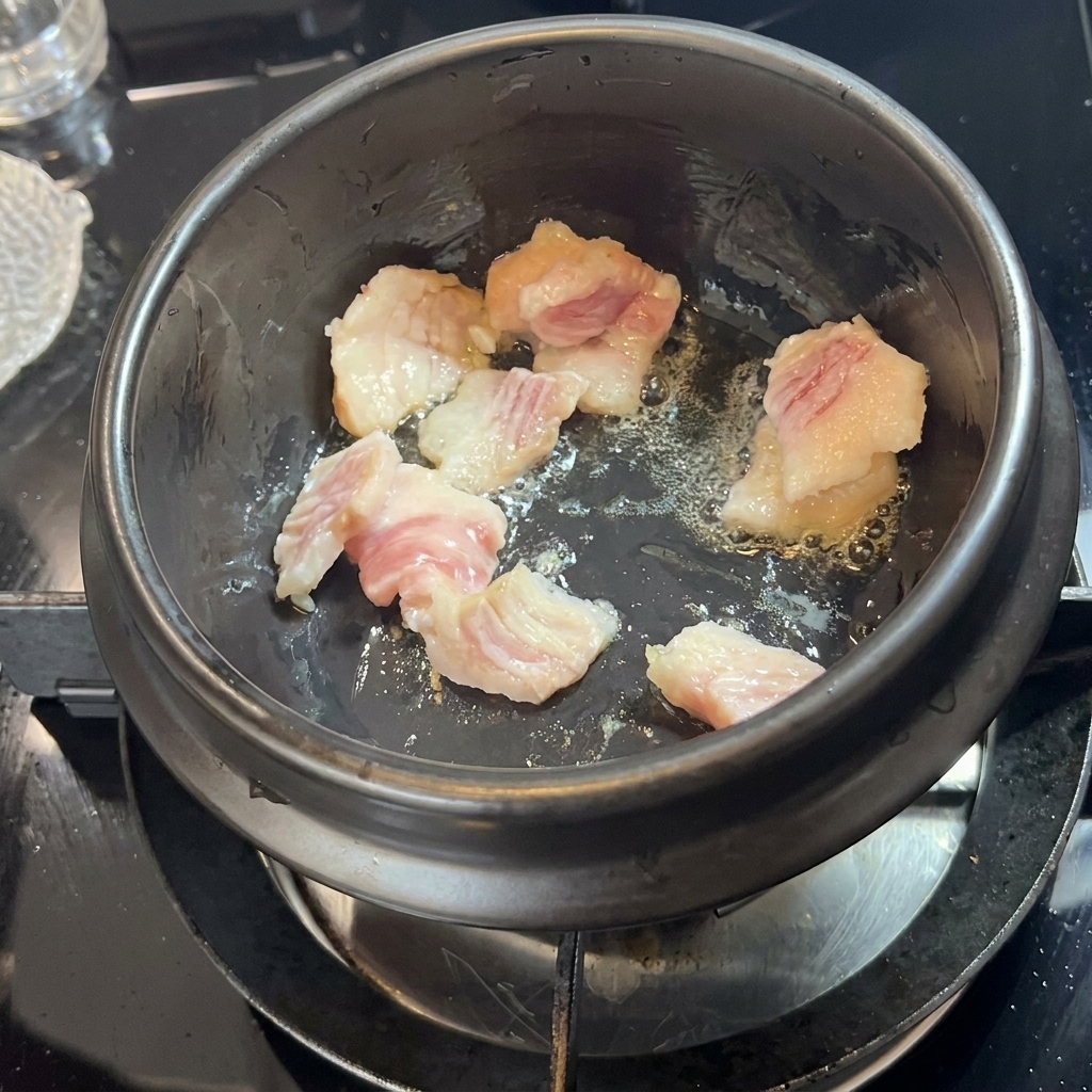 Trozos de panceta sofriéndose y soltando grasa en una olla de piedra negra.