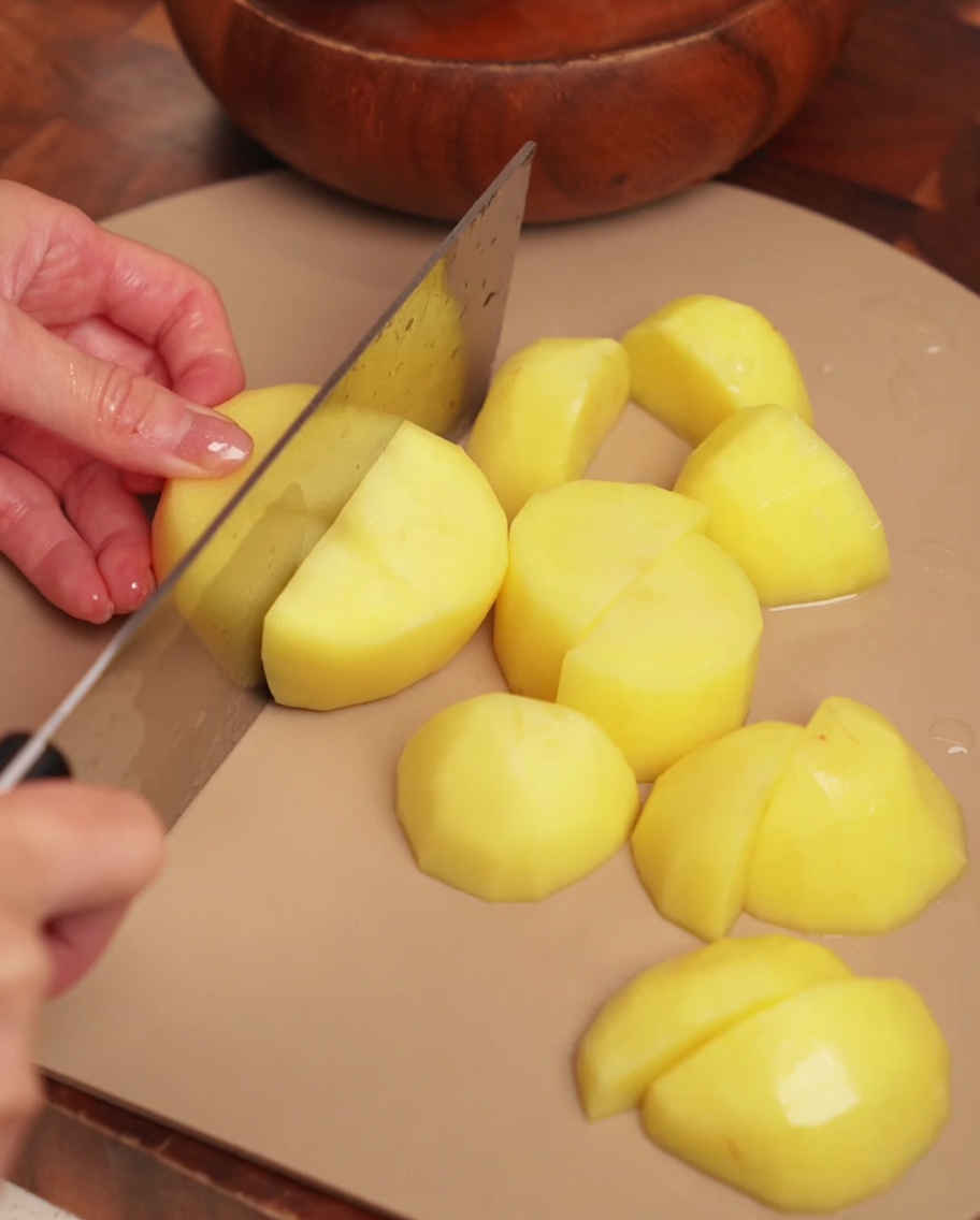 Patatas peladas siendo cortadas en trozos grandes y uniformes sobre una tabla de cortar de madera.