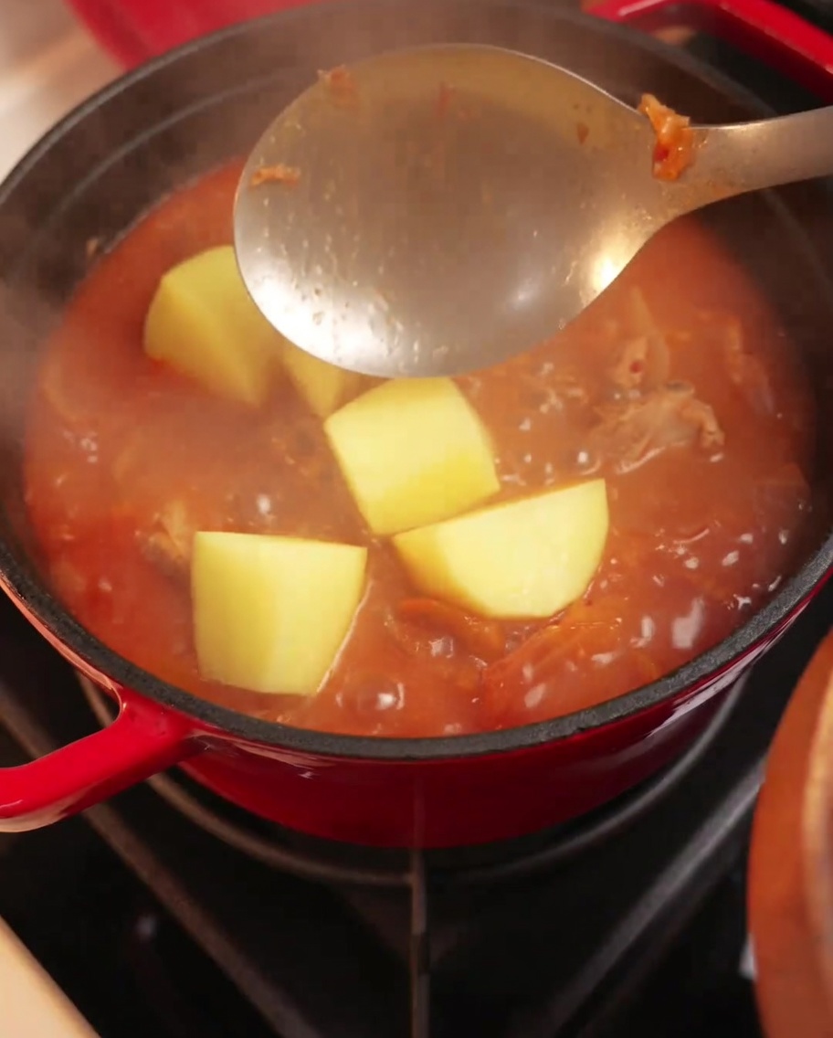 Trozos grandes de patata siendo añadidos a una olla burbujeante de estofado de cerdo y kimchi.