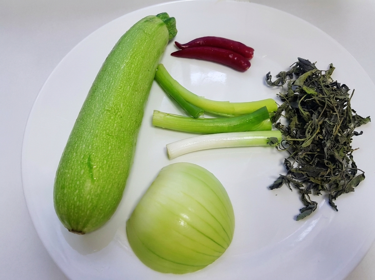 Ingredientes crudos, incluyendo una calabaza verde entera, cebollines, cebolla blanca, hojas de perilla silvestres y chiles rojos exhibidos en un plato blanco.