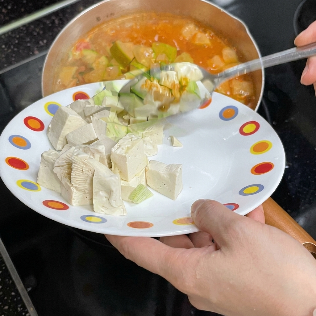 Una persona deslizando cubos de tofu blanco desde un plato colorido a una olla de estofado rojo hirviendo.