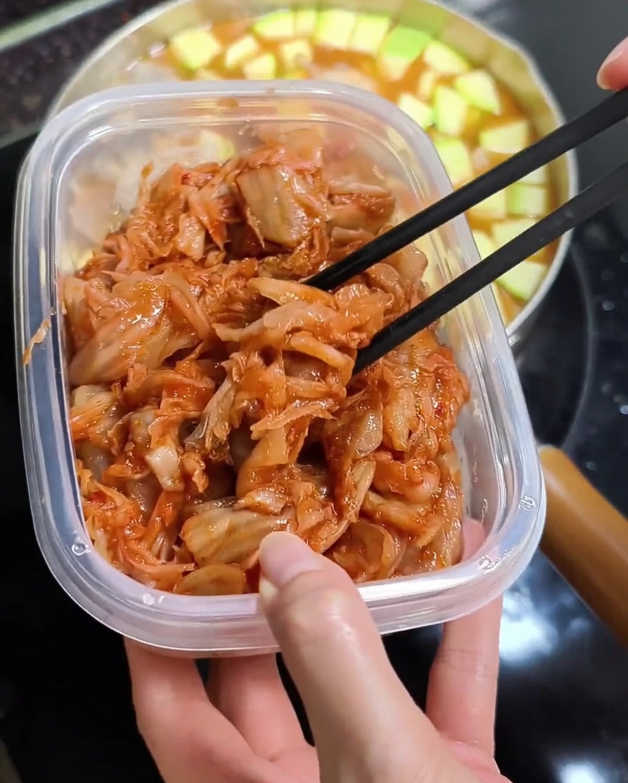 Palillos levantando kimchi picado de color rojo brillante desde un recipiente de plástico sobre una olla de estofado caliente.