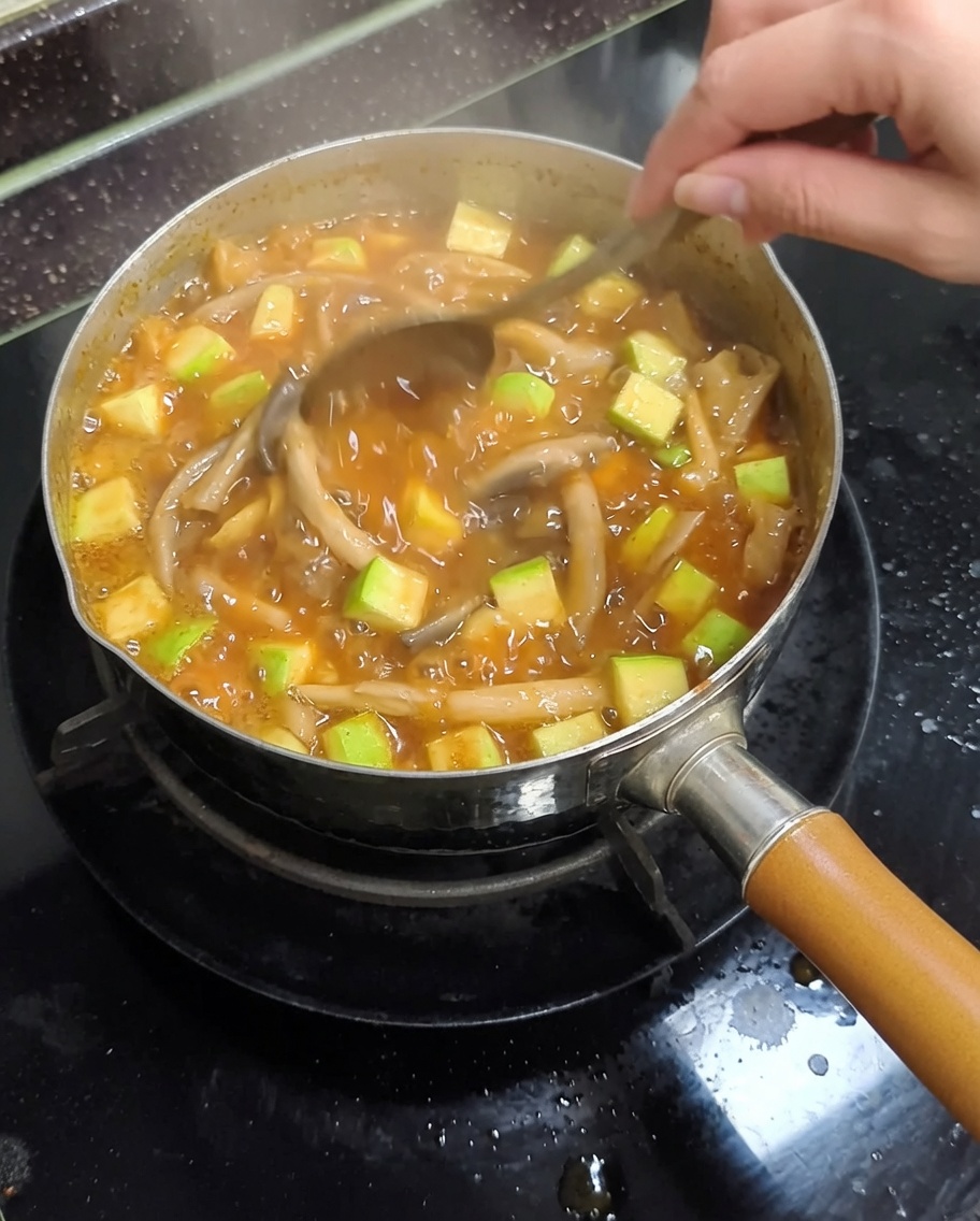 Una olla de metal en los fogones con estofado de pasta de soja hirviendo siendo removido con una cuchara de metal, mostrando calabacines y setas.