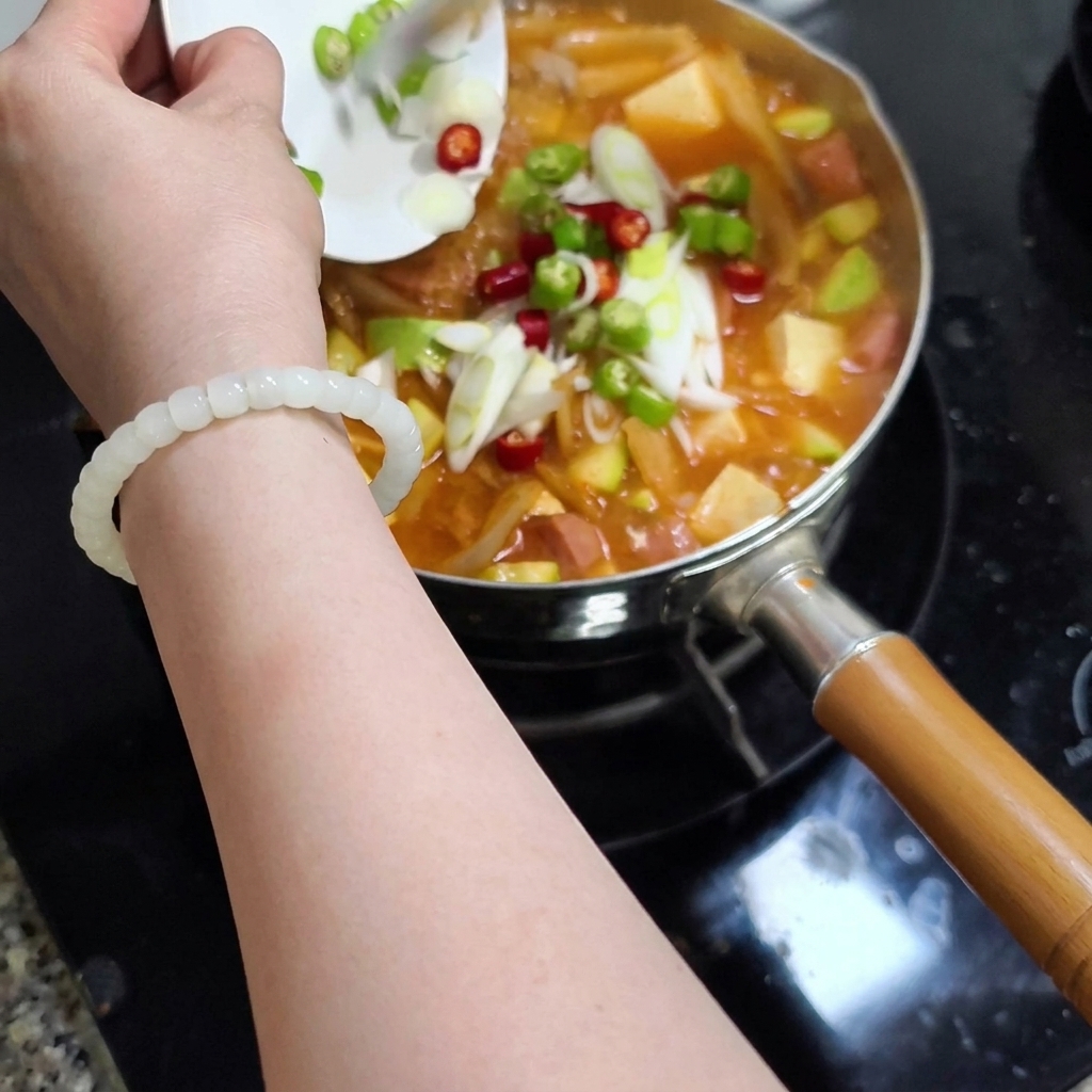 Una mano sosteniendo un pequeño plato blanco deslizando chiles verdes y rojos picados y cebolletas en rodajas a una olla de estofado burbujeante.