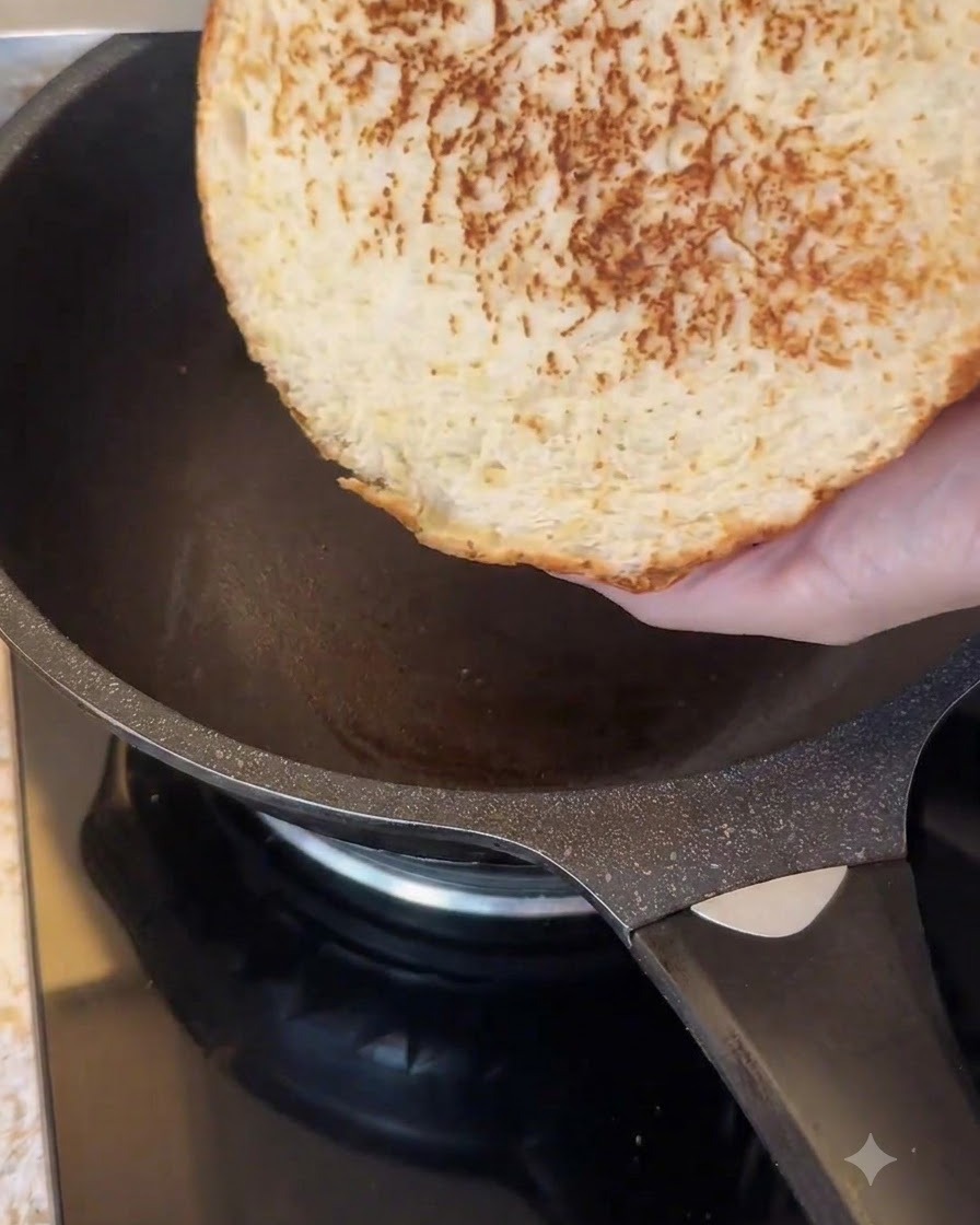El lado cortado de un gran pan de hamburguesa tostándose en una sartén oscura antiadherente.