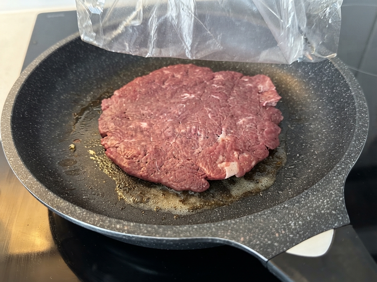 Una gran hamburguesa de carne cruda cocinándose en una sartén antiadherente engrasada sobre el fogón.