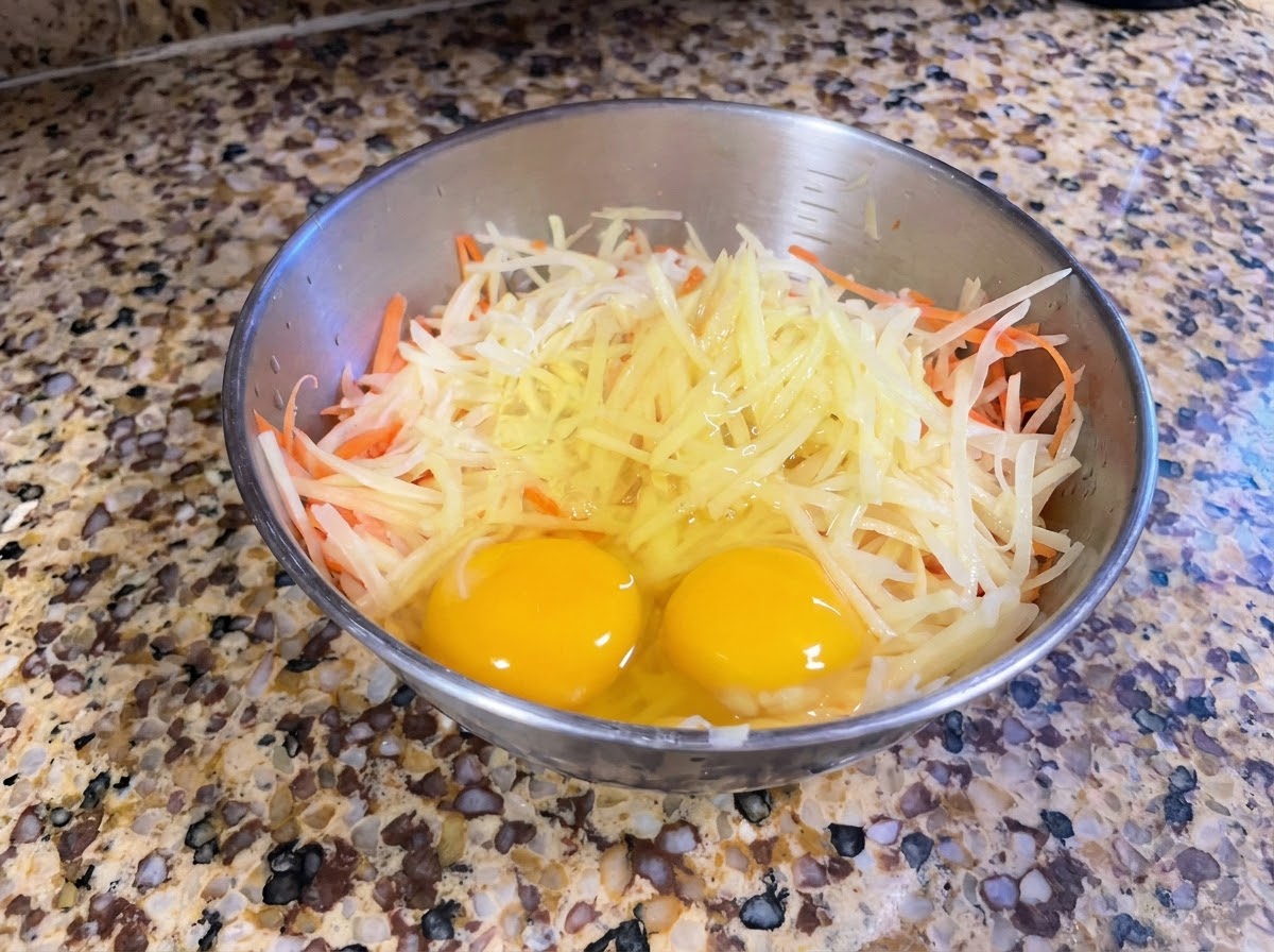 Un bol de metal que contiene patatas y zanahorias recién ralladas con dos huevos enteros encima.
