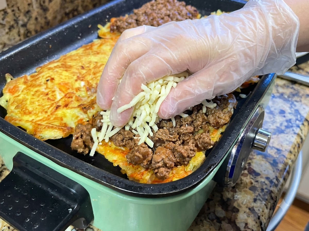 Manos con guantes protectores colocando un puñado de queso mozzarella rallado sobre una capa de salsa de carne cocida extendida en una tortilla de patata a la sartén.