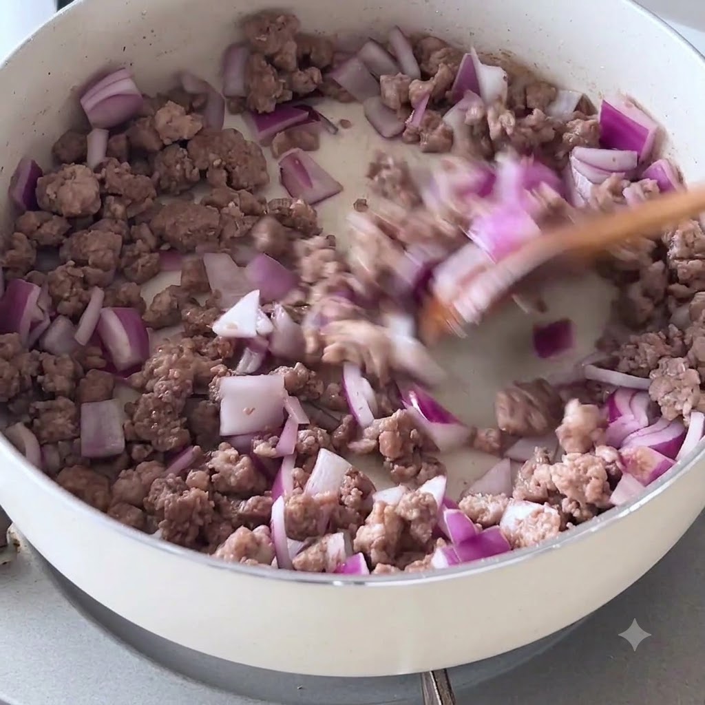 Cebolla morada picada y carne picada dorada removiéndose con una espátula de madera en una sartén blanca.
