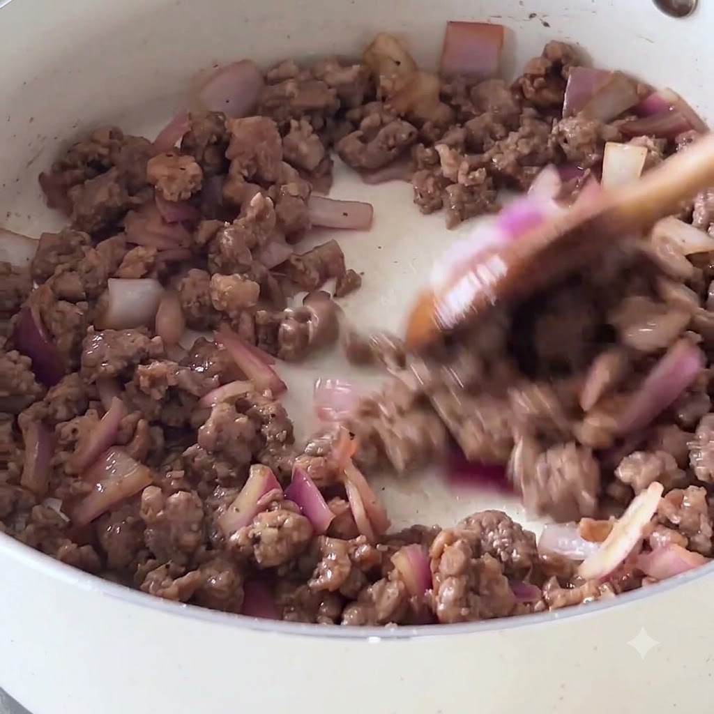 Salteado de carne picada y cebolla morada en una sartén blanca antiadherente usando una cuchara de madera.