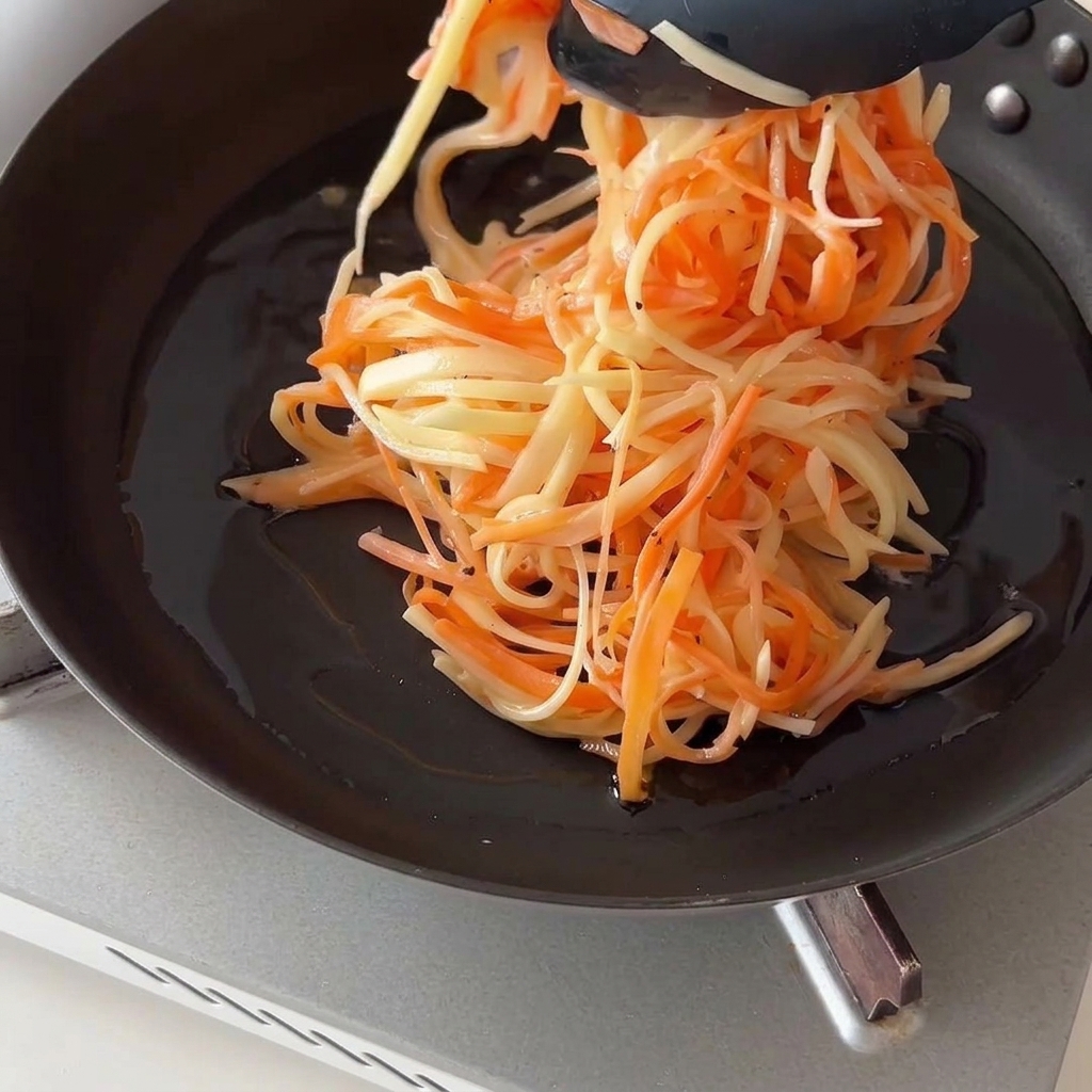 Usando unas pinzas de cocina negras para colocar una montaña de patatas y zanahorias ralladas en una sartén aceitada.