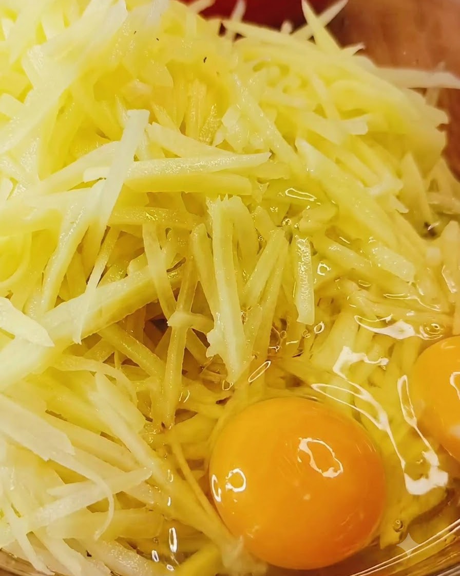 Huevos enteros cascados directamente sobre un tazón de patatas ralladas crudas y condimentos.
