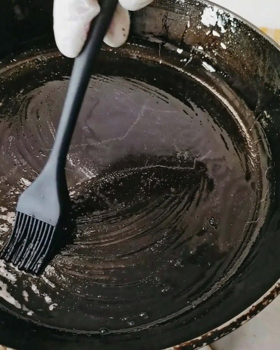 Una sartén negra siendo cepillada con aceite usando una brocha de repostería de silicona negra.