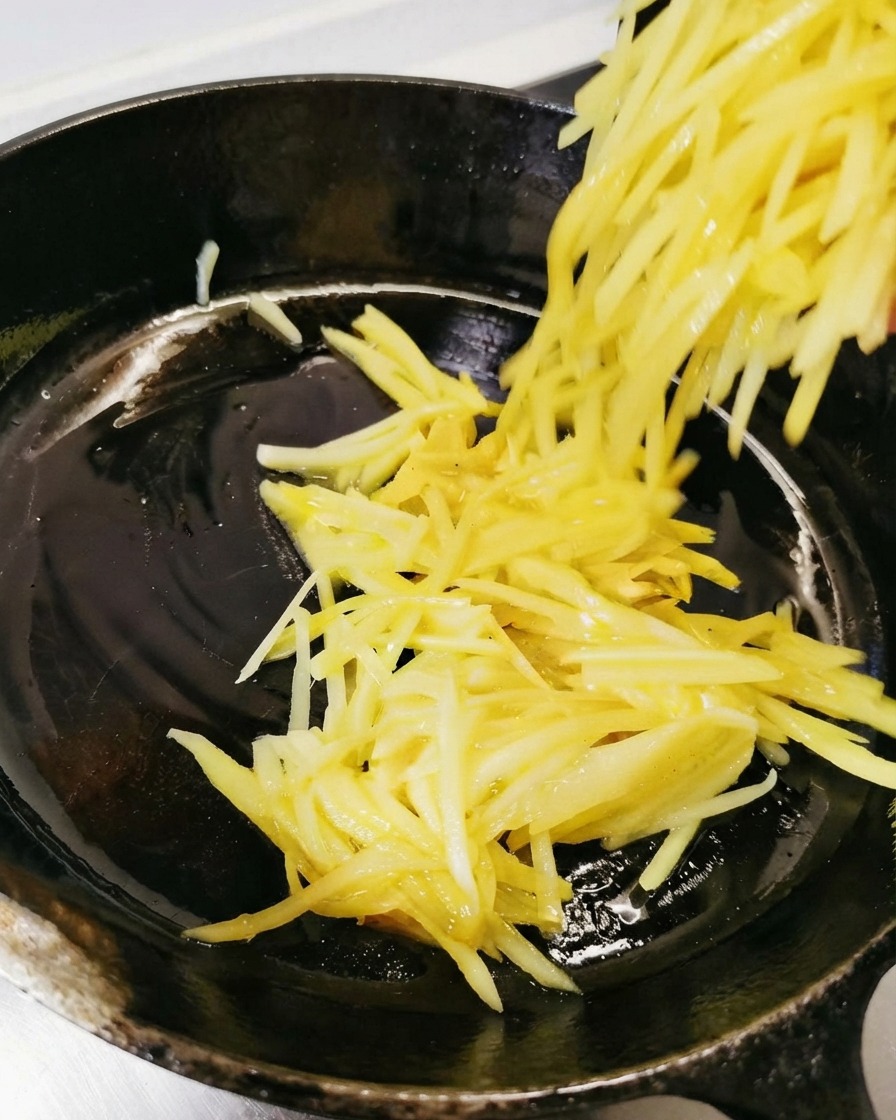 Patatas ralladas siendo vertidas y extendidas en la sartén negra engrasada.