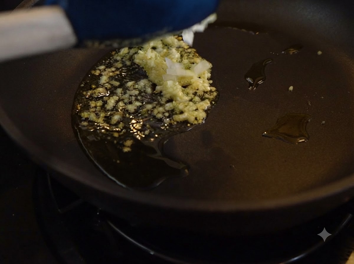 Ajo picado y cebollas sofriéndose en aceite dentro de una sartén oscura.