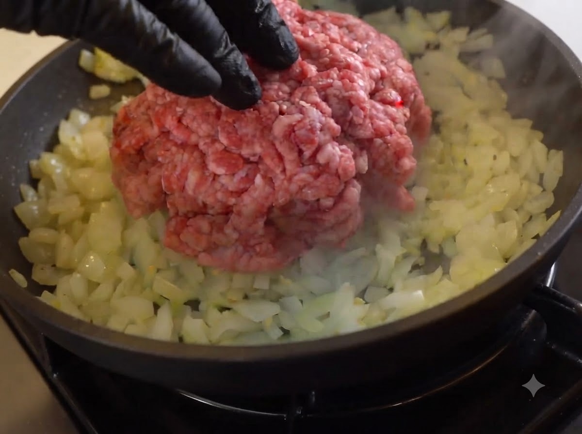 Una porción de carne picada cruda colocada sobre cebollas sofritas en una sartén.