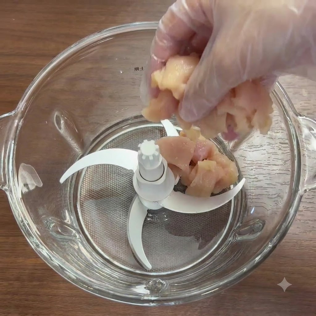 Trozos de pechuga de pollo y verduras frescas dentro del vaso de una batidora de vidrio con cuchillas.