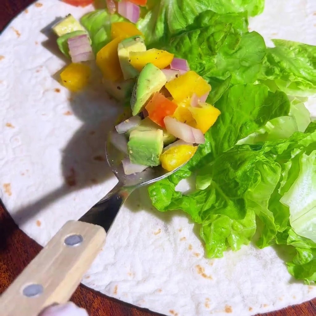 Una cuchara colocando la salsa de aguacate y vegetales sobre la lechuga en la tortilla.