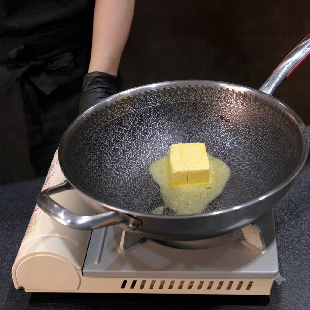 Un bloque de mantequilla amarilla derritiéndose en un wok de metal, revuelto con un pequeño batidor sobre el fuego.