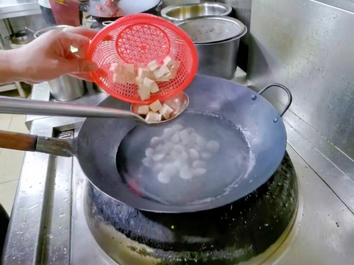 Una mano sosteniendo una cesta colador roja, dejando caer tofu en dados en un wok con agua y almejas.