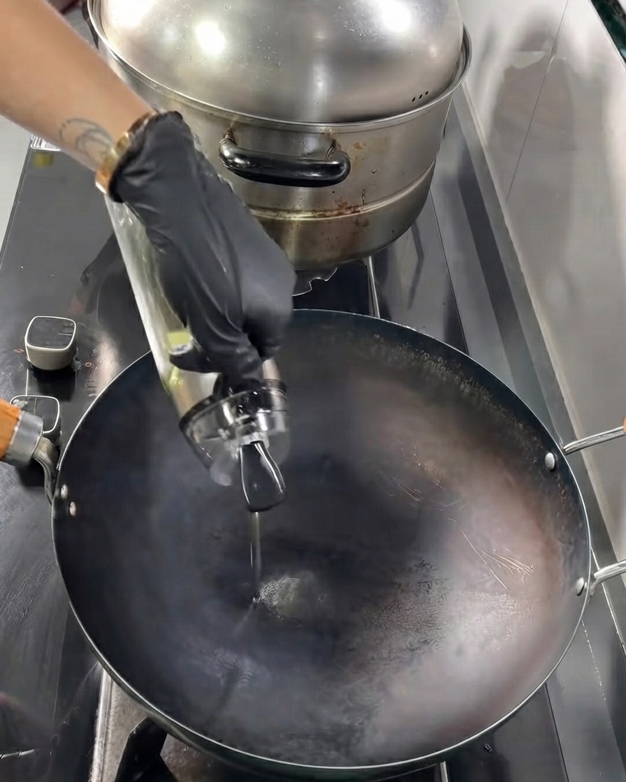 Aceite de cocina cayendo desde una botella de vidrio hacia un wok negro caliente.