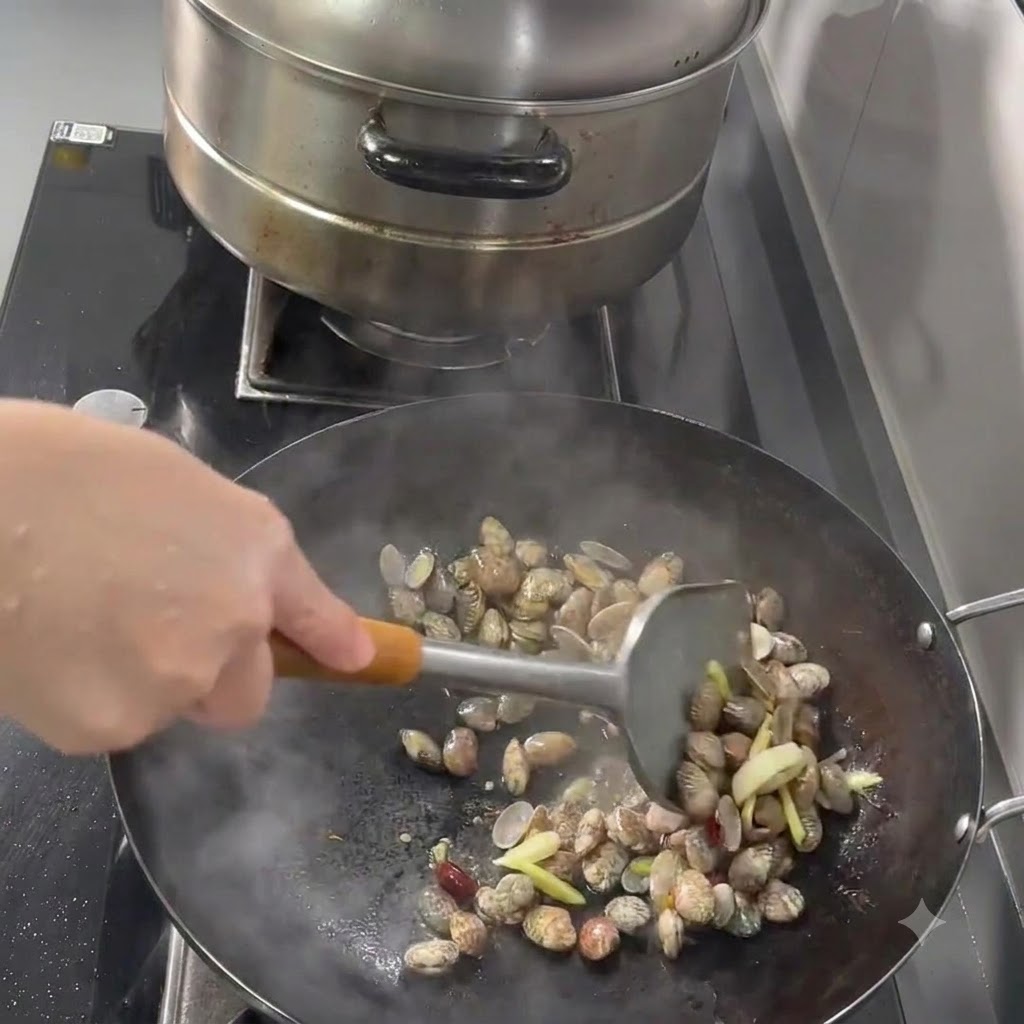 Almejas, ajo, jengibre y chiles salteándose en un wok con una espátula de metal.
