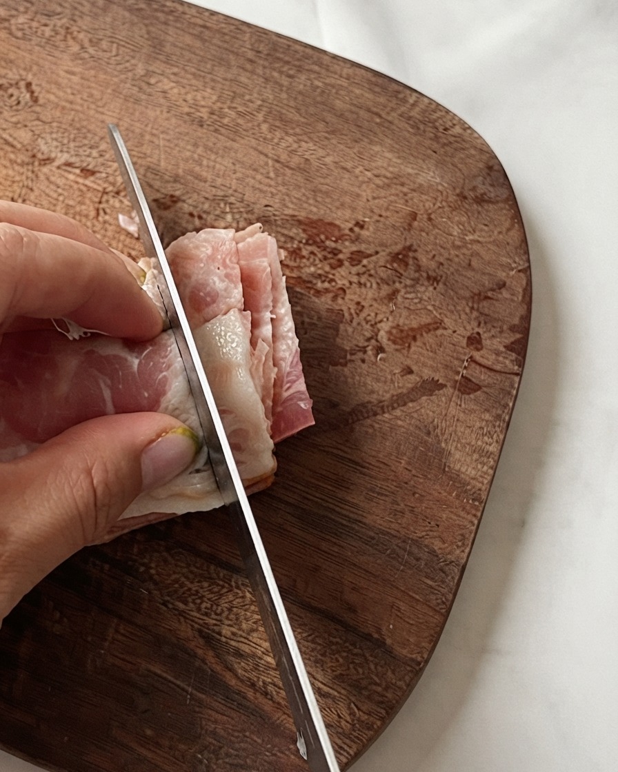 La mano de un chef sosteniendo tiras de beicon apiladas mientras un cuchillo las corta en trozos pequeños sobre una tabla de madera.