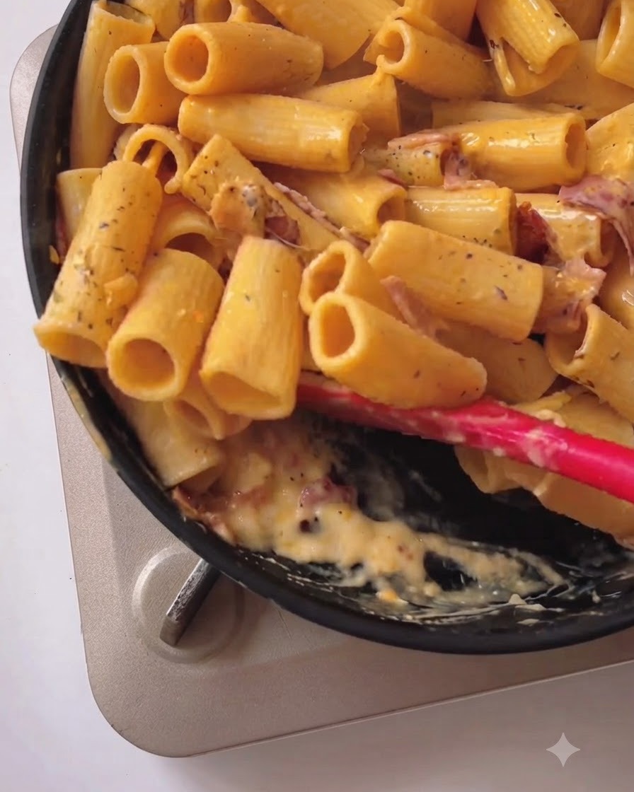 Pasta rigatoni siendo revuelta y mezclada con salsa cremosa amarilla y beicon en una sartén negra usando una espátula de silicona roja.