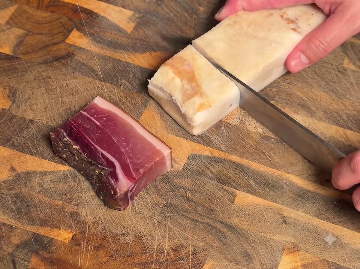 Un cuchillo cortando un bloque de carne curada y tocino en una tabla de cortar de madera texturizada.