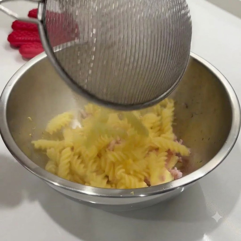 Pasta en espiral recién cocida vertiéndose desde un colador de alambre a un bol de acero inoxidable con los ingredientes.