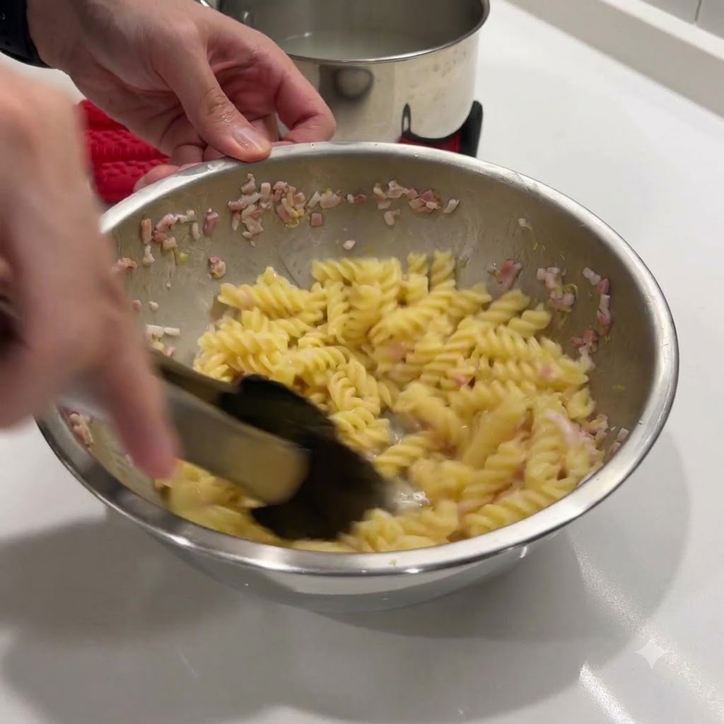 Removiendo la pasta caliente y la mezcla de mantequilla en un bol de metal con unas pinzas negras.