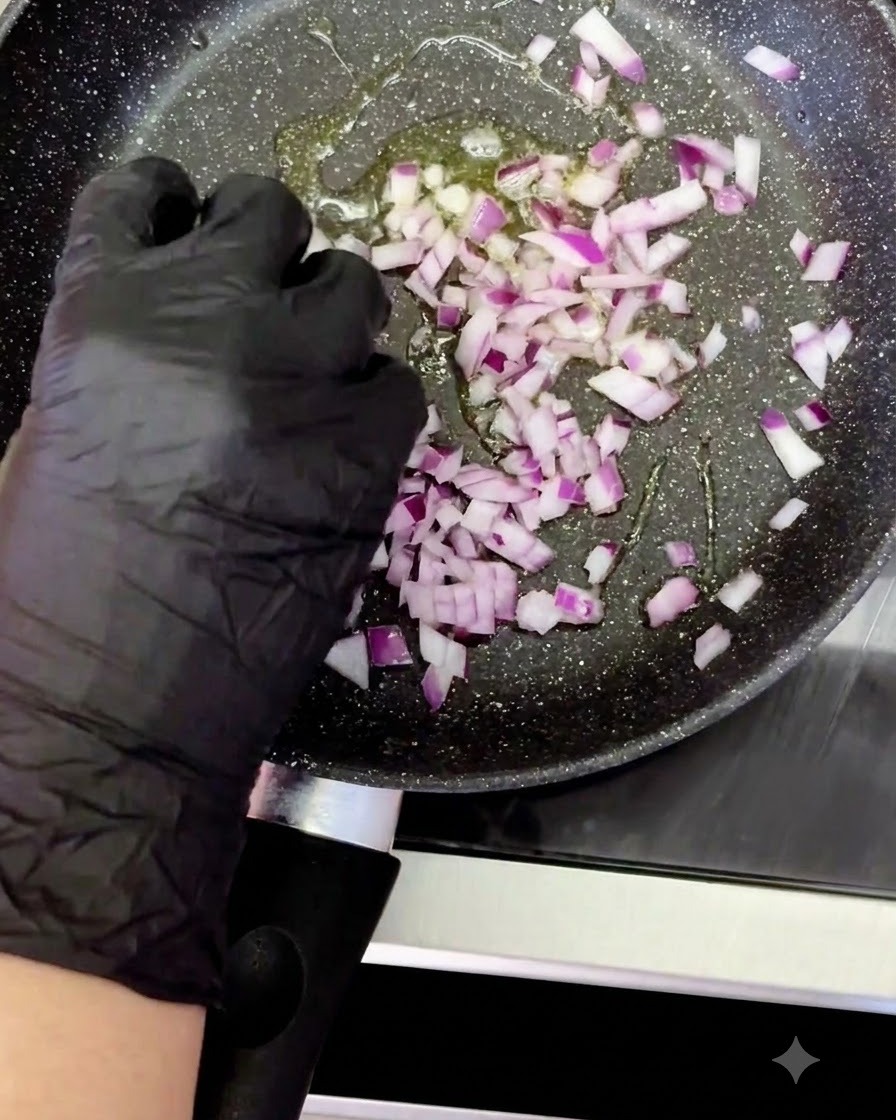 Una mano con guantes agregando cebolla morada en cubos a una sartén caliente con aceite chisporroteante.