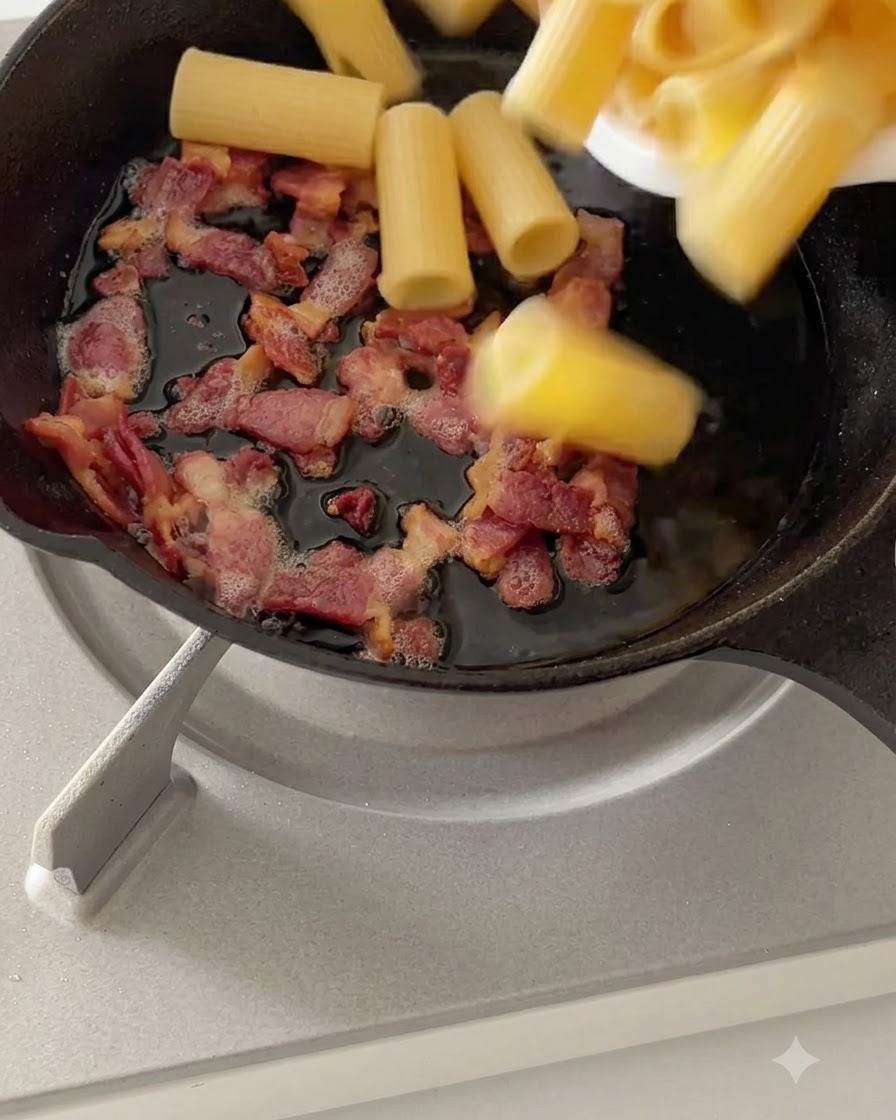 Pasta rigatoni hervida mezclándose con el beicon cocinado en una sartén a fuego lento.