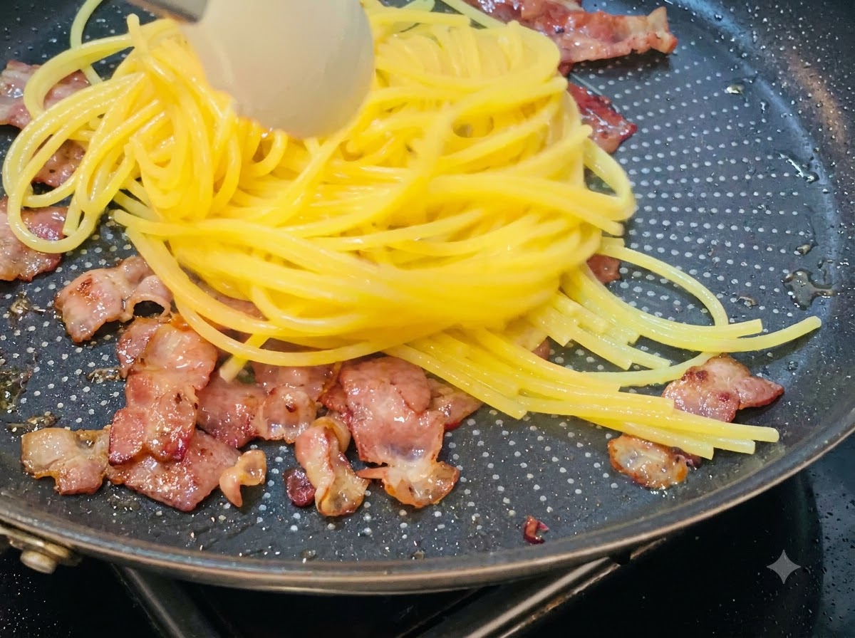 Espagueti cocido siendo levantado y transferido a una sartén que contiene trozos de tocino frito.