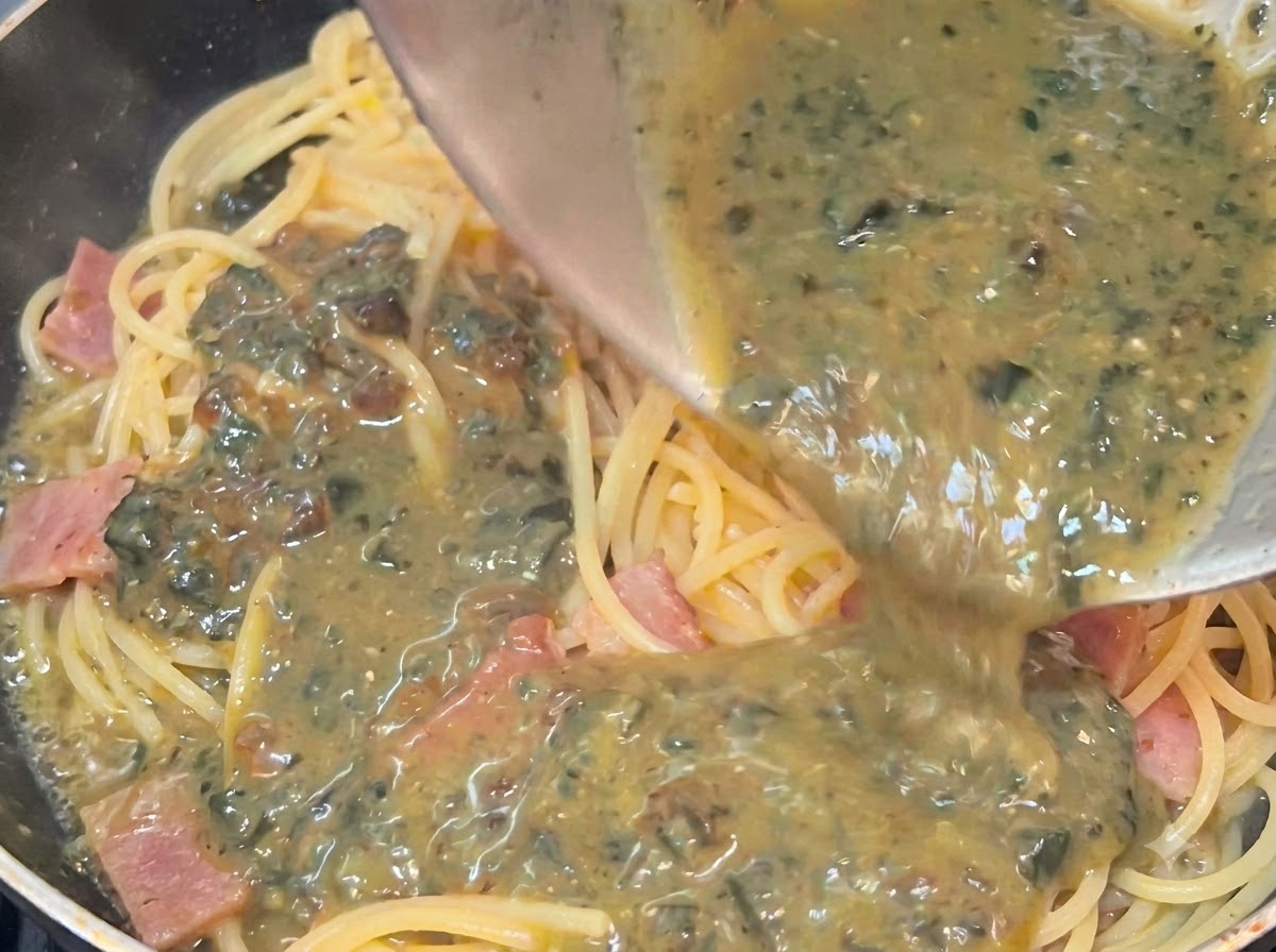 Salsa de huevo centenario cremosa y salpicada siendo vertida desde un tazón sobre el espagueti caliente y el tocino en una sartén.