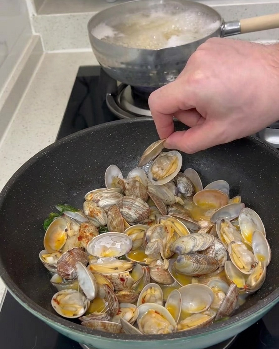 La mano de una persona recogiendo una concha de almeja abierta de una sartén llena de almejas cocidas y salsa.