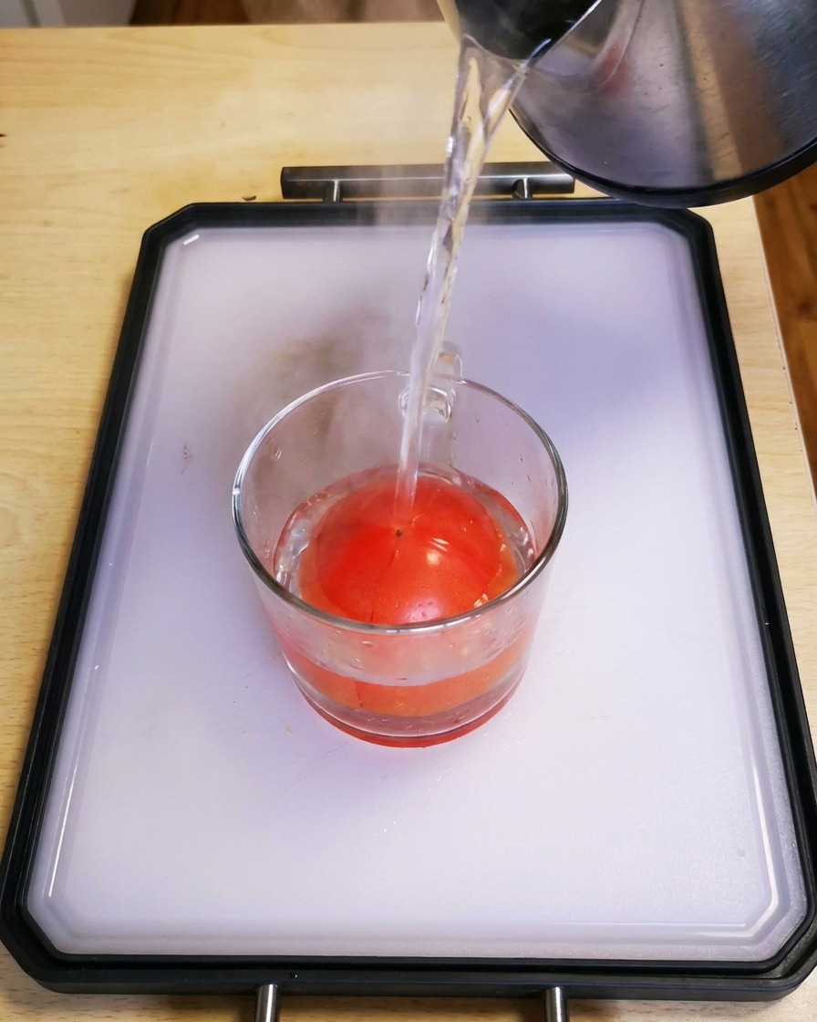 Agua caliente vertiéndose desde un hervidor en una taza de cristal que contiene un tomate rojo fresco.